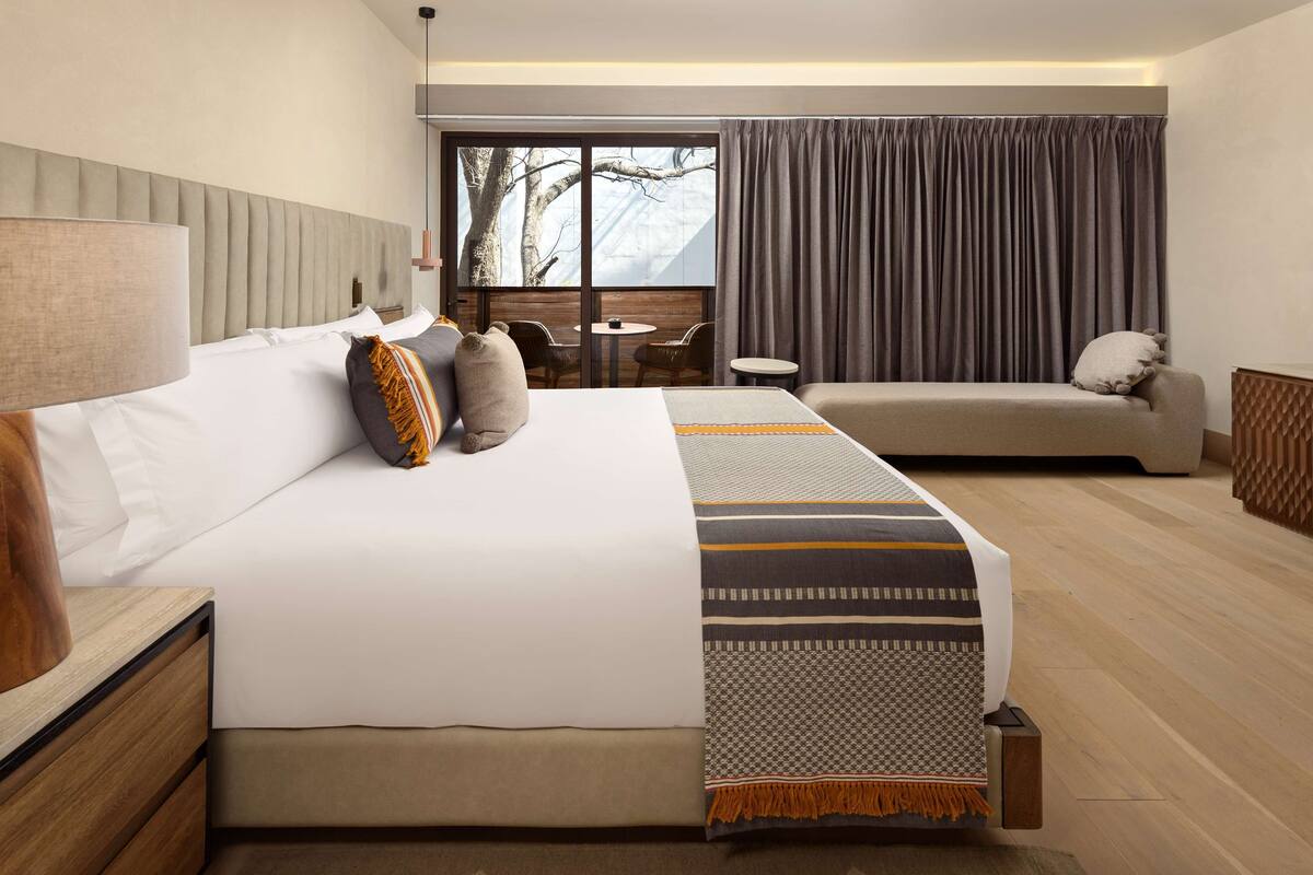 A modern hotel room with a large bed, a sofa, a small table, and a view of the outdoors.