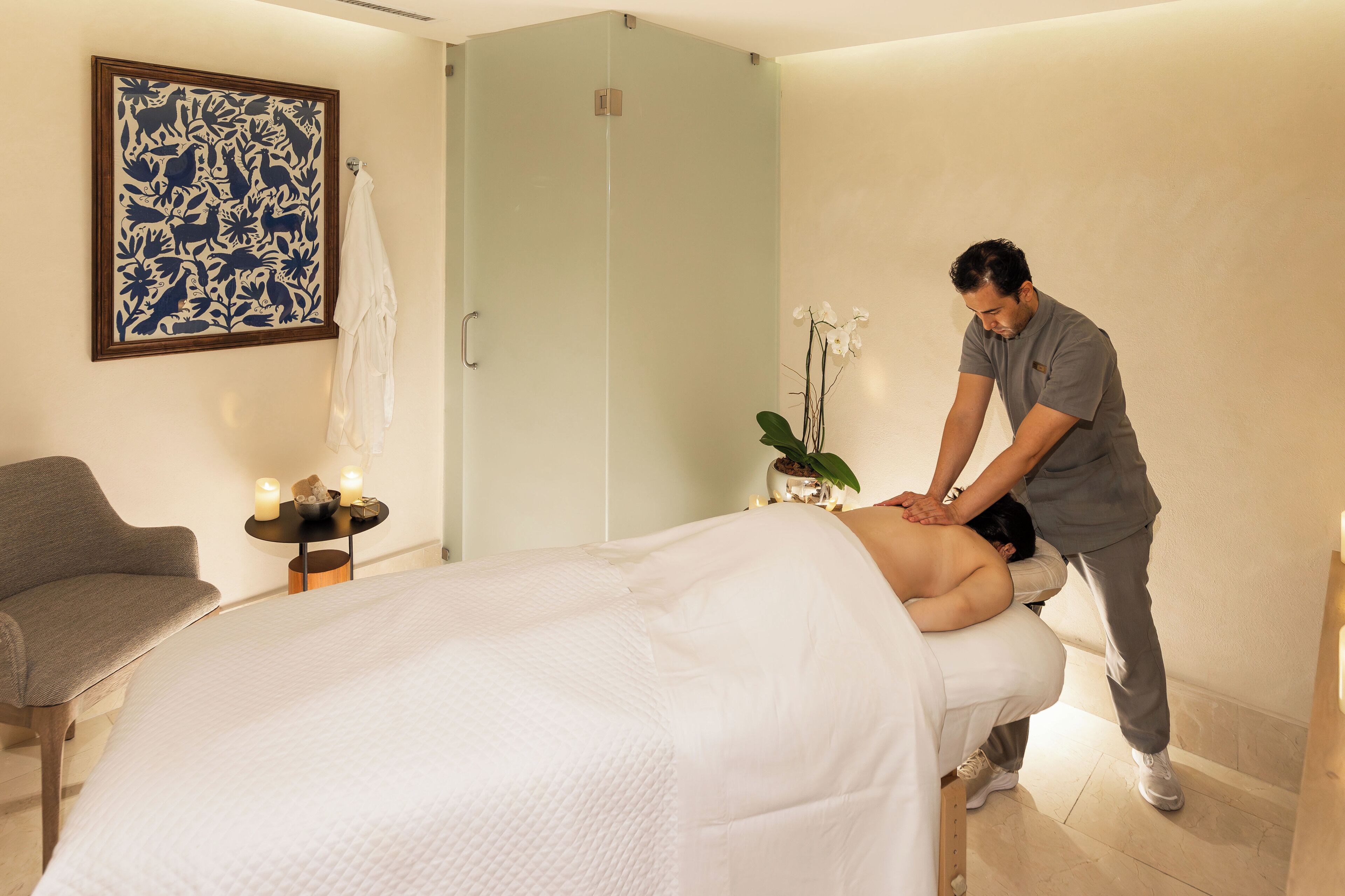A massage therapy room with a bed, a massage table, a chair, a small table with candles, and a potted plant.
