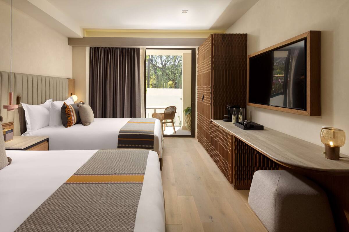 A hotel room with two beds, a TV, a desk, and a view of the outdoors.