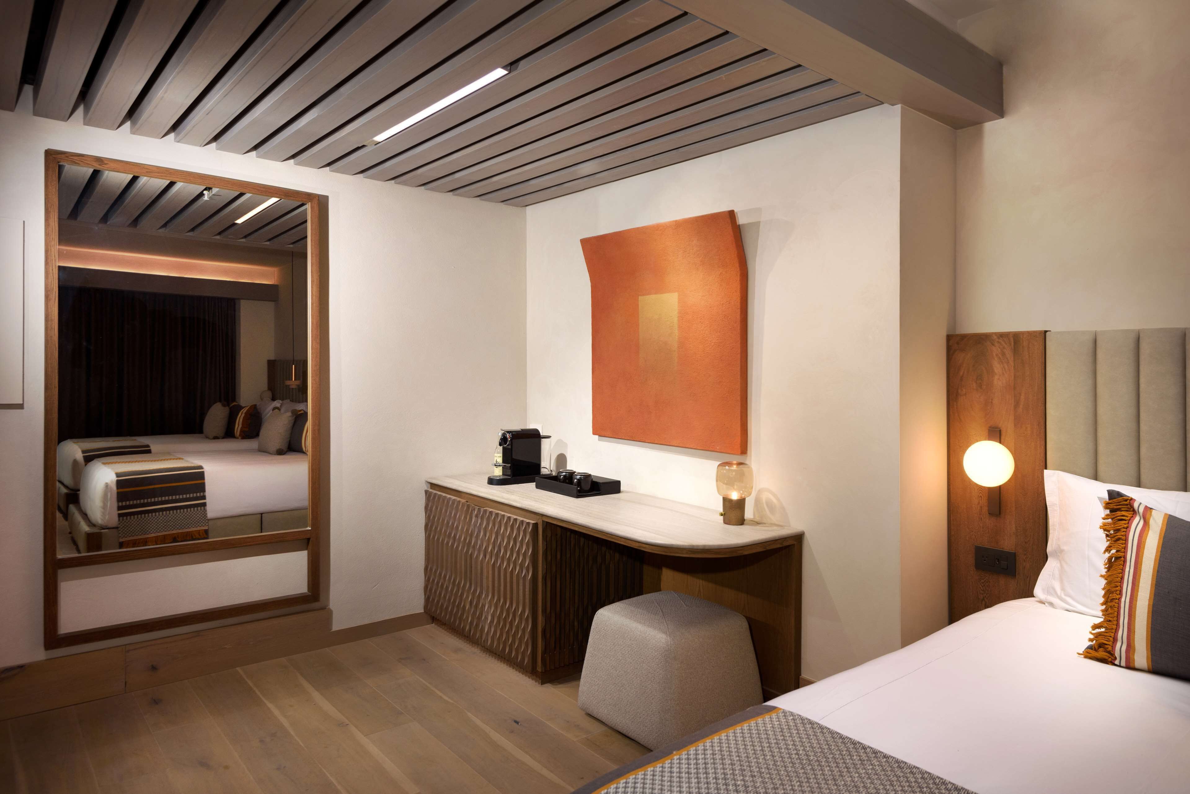 A modern hotel room with a wooden ceiling, a bed with a headboard, a bedside table with a lamp, and a large mirror.