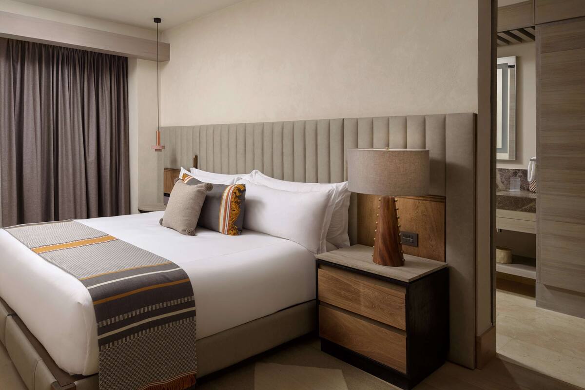 A modern hotel room with a large bed, a nightstand with a lamp, and a view of the hallway.