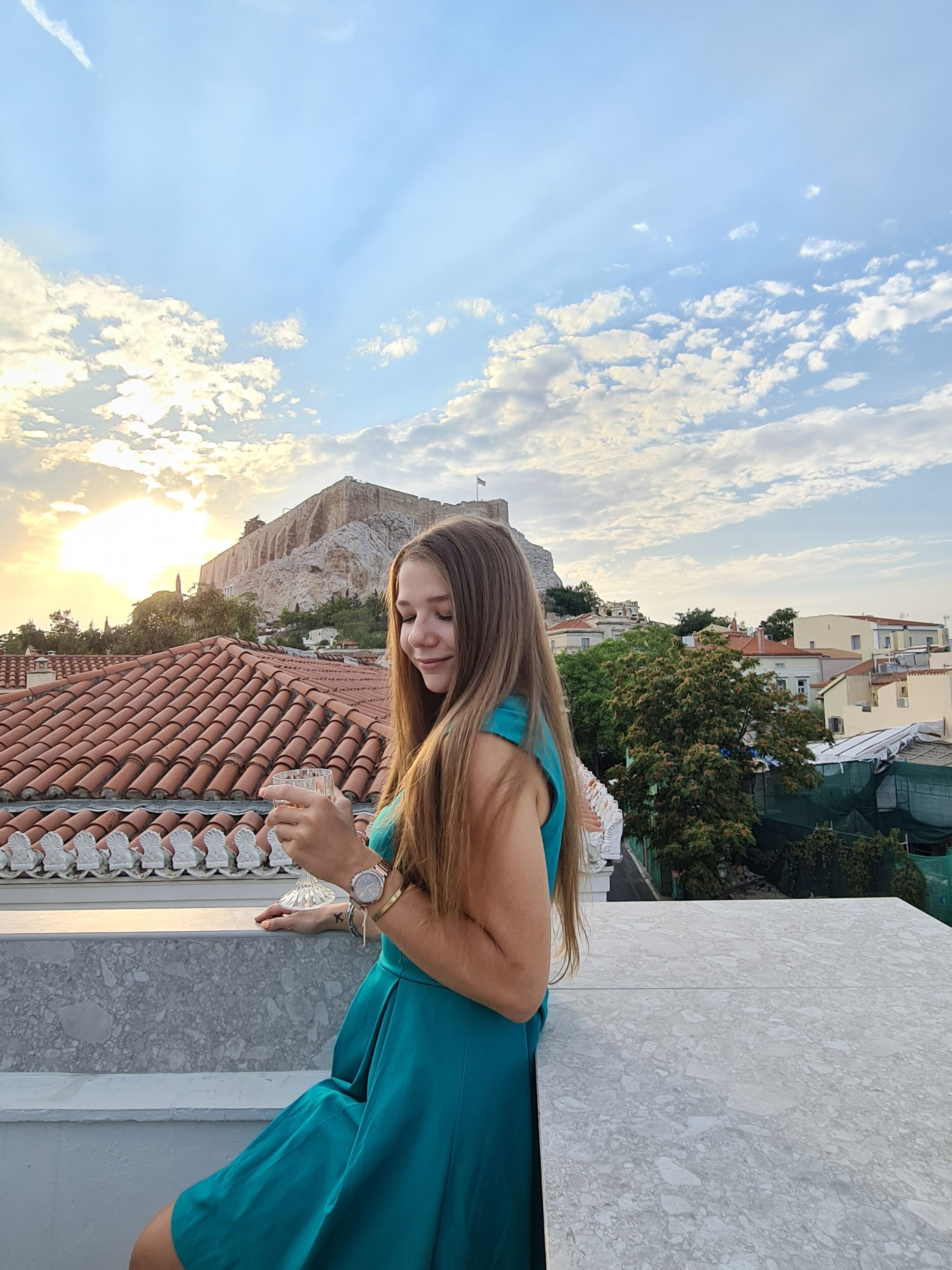 A woman in a teal dress on a rooftop with terracotta tiles, overlooking a cityscape with a prominent hill and buildings in the background.