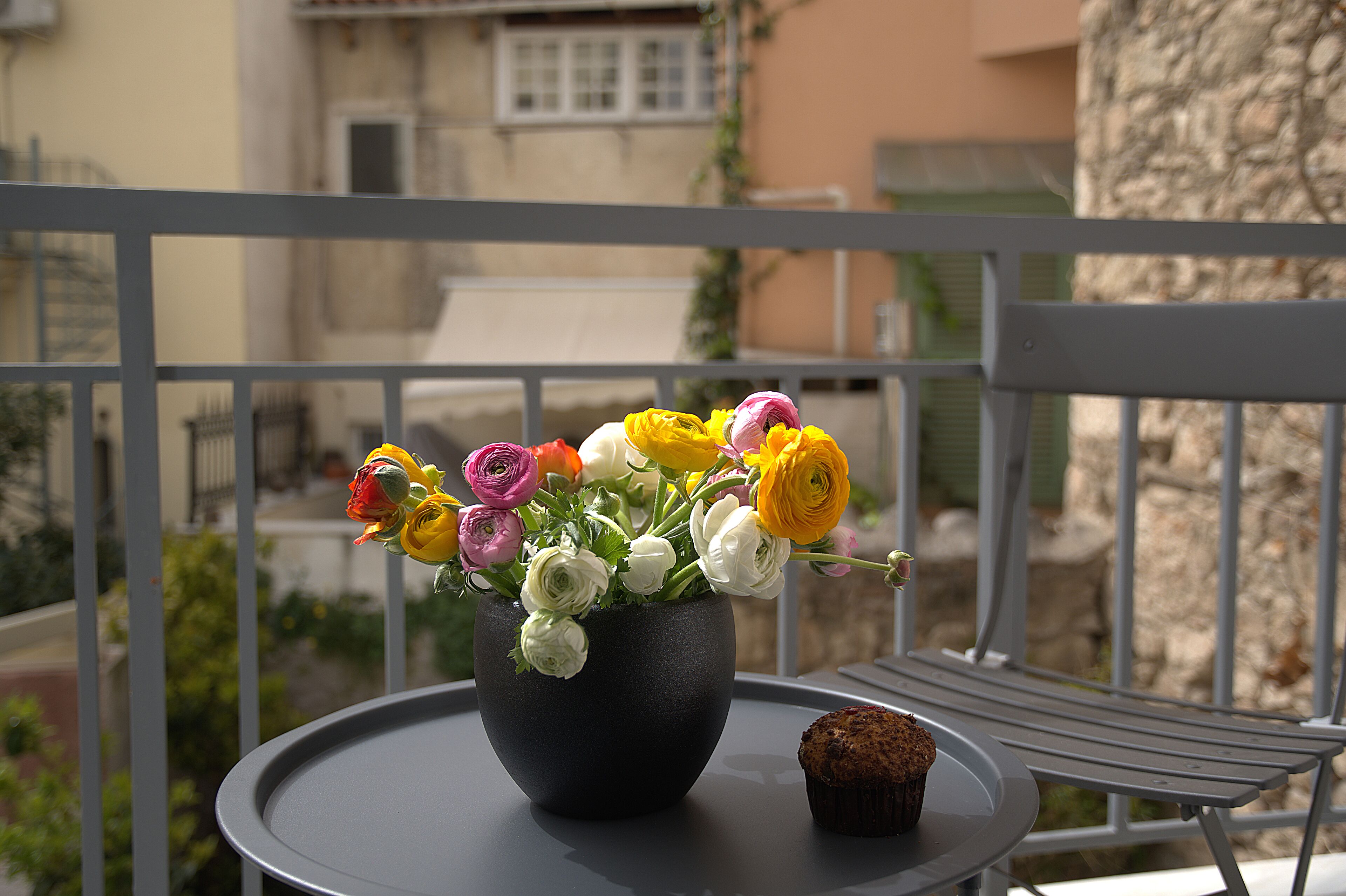 Un balcon avec une table ronde, un vase noir orné de fleurs et un muffin.