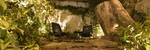 An outdoor seating area with two wicker chairs and a table, surrounded by lush greenery and a large tree trunk. - CityZoe Athens Suites (Athens)