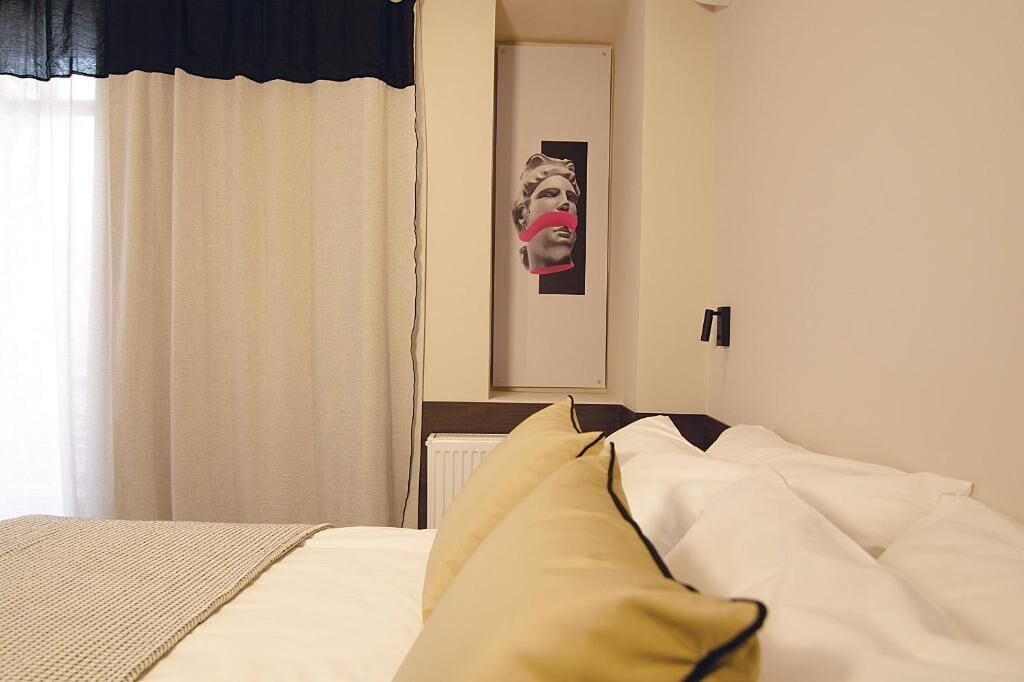 A hotel room with a bed, a window with curtains, a radiator, and a wall-mounted artwork.