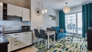 Eine moderne Wohnung mit Küche, Essbereich und Wohnzimmer. Sie verfügt über einen Streifen Teppich, blaue Vorhänge und ein großes Fenster mit Blick auf Gebäude.
