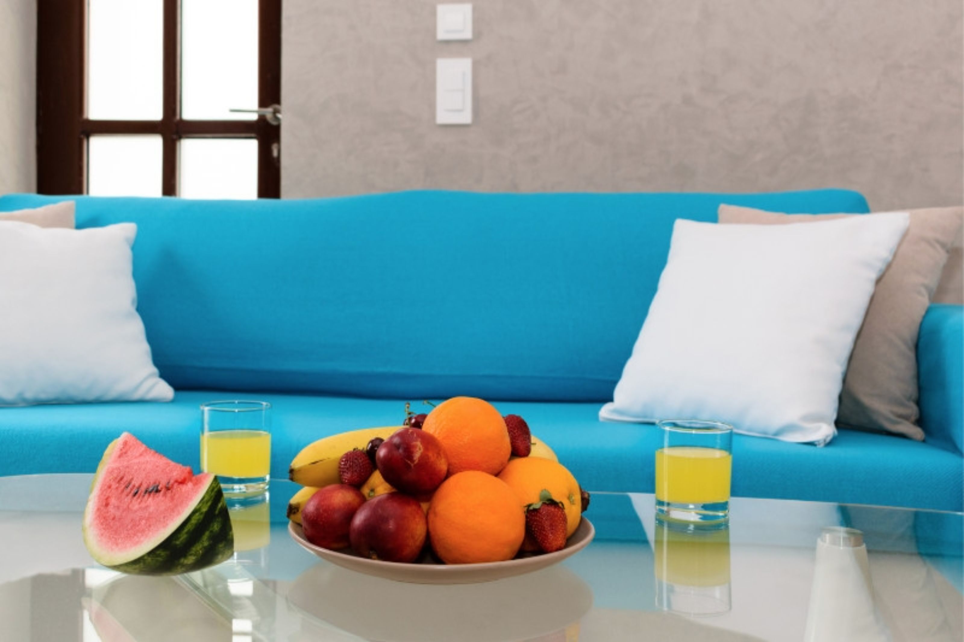 A room with a blue sofa, a glass table with a fruit bowl, and two glasses of orange juice.