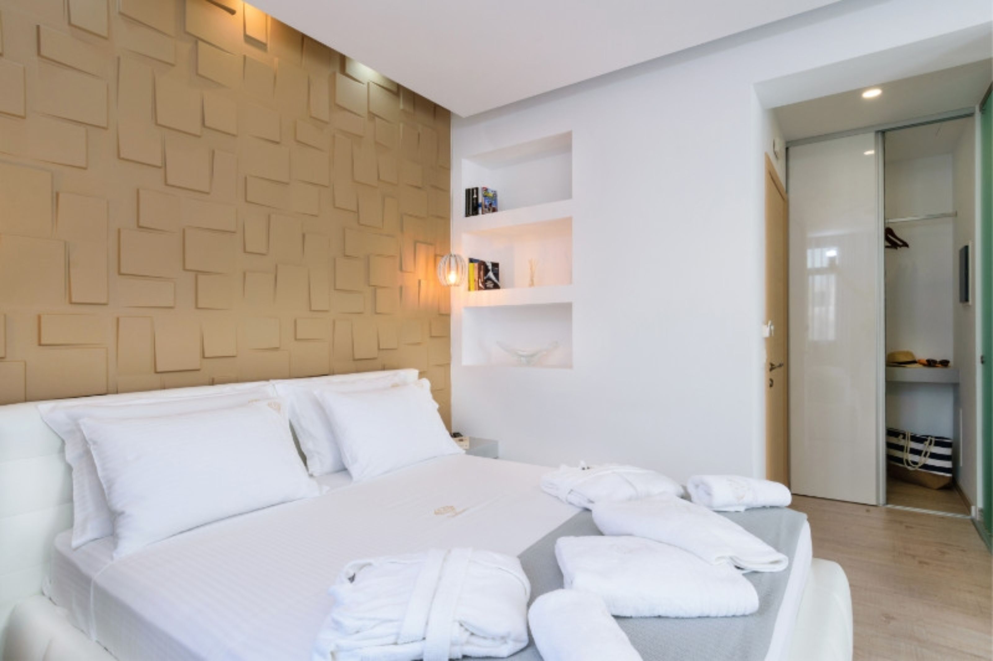 Una habitación de hotel moderna con una cama grande, ropa de cama blanca, un panel de madera decorativo, una estantería empotrada con libros y un vestidor.