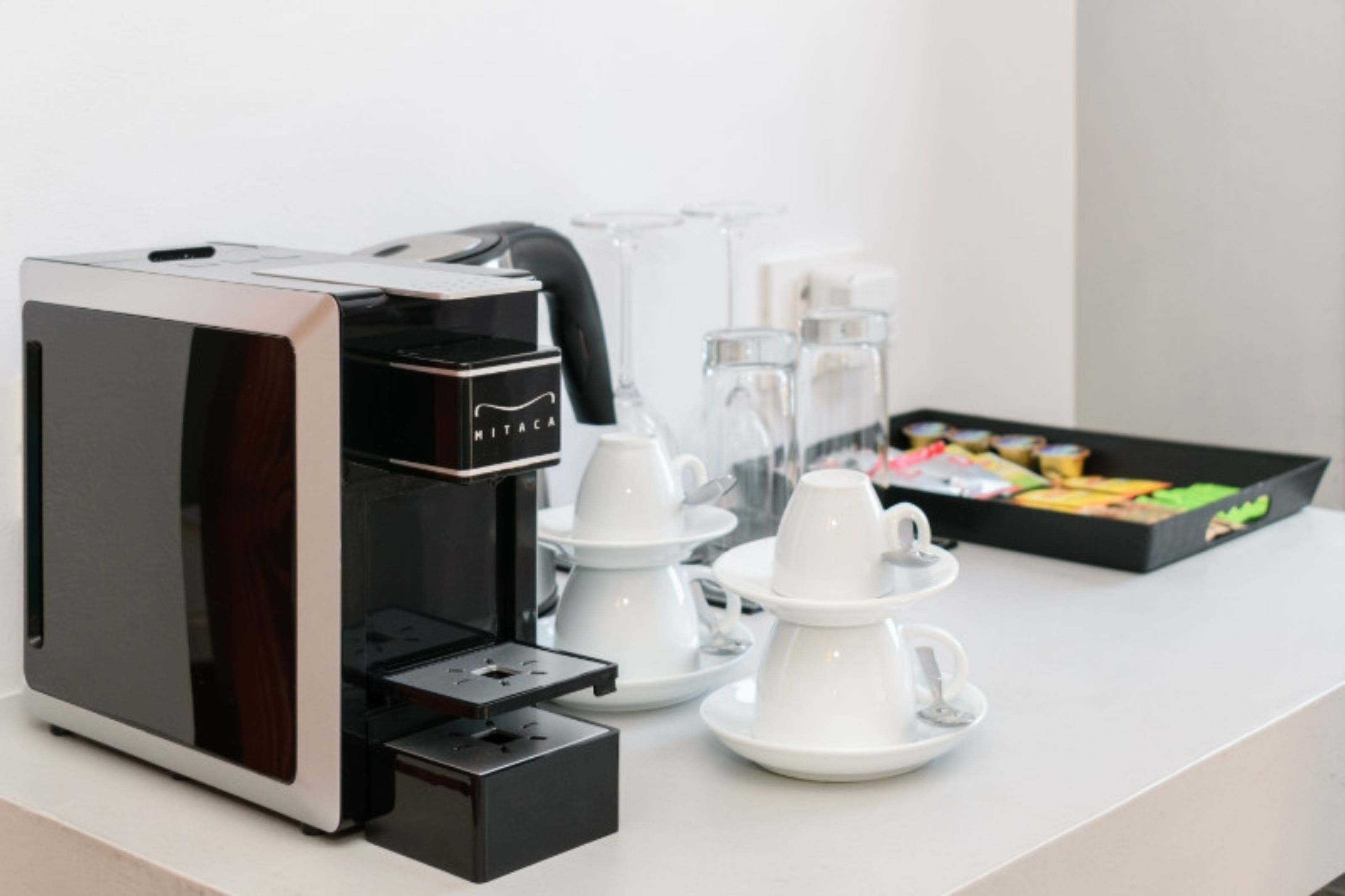 A coffee machine with a brand name, a tray of white cups, and a box of colorful cookies on a white surface.