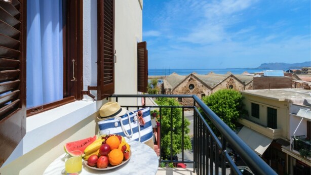 Een balkon met een tafel vol fruit en een strohoed, met uitzicht op een kustplaatsje met traditionele gebouwen.