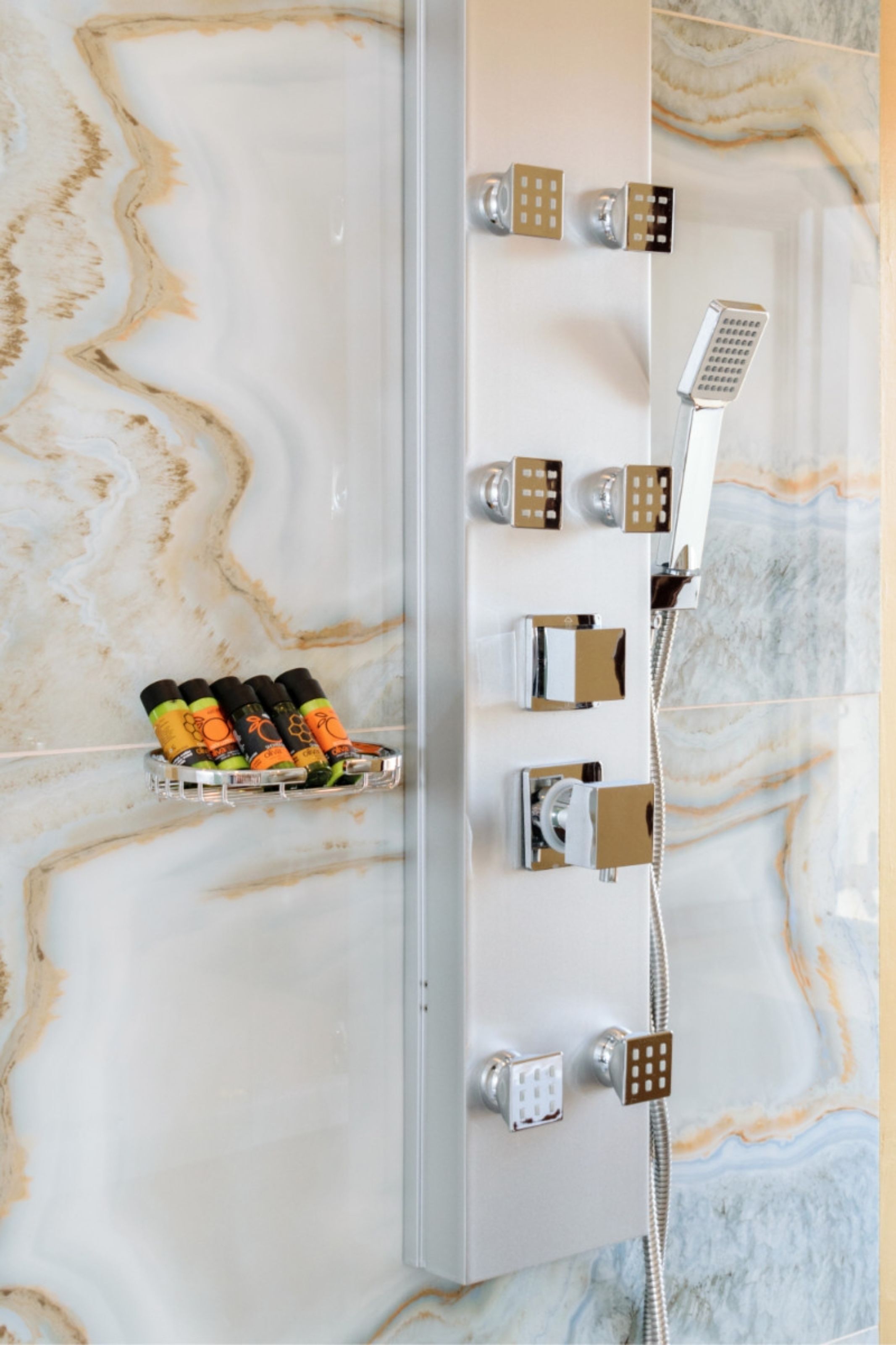 A bathroom with a marble wall, a shower with multiple controls, and a soap dispenser.