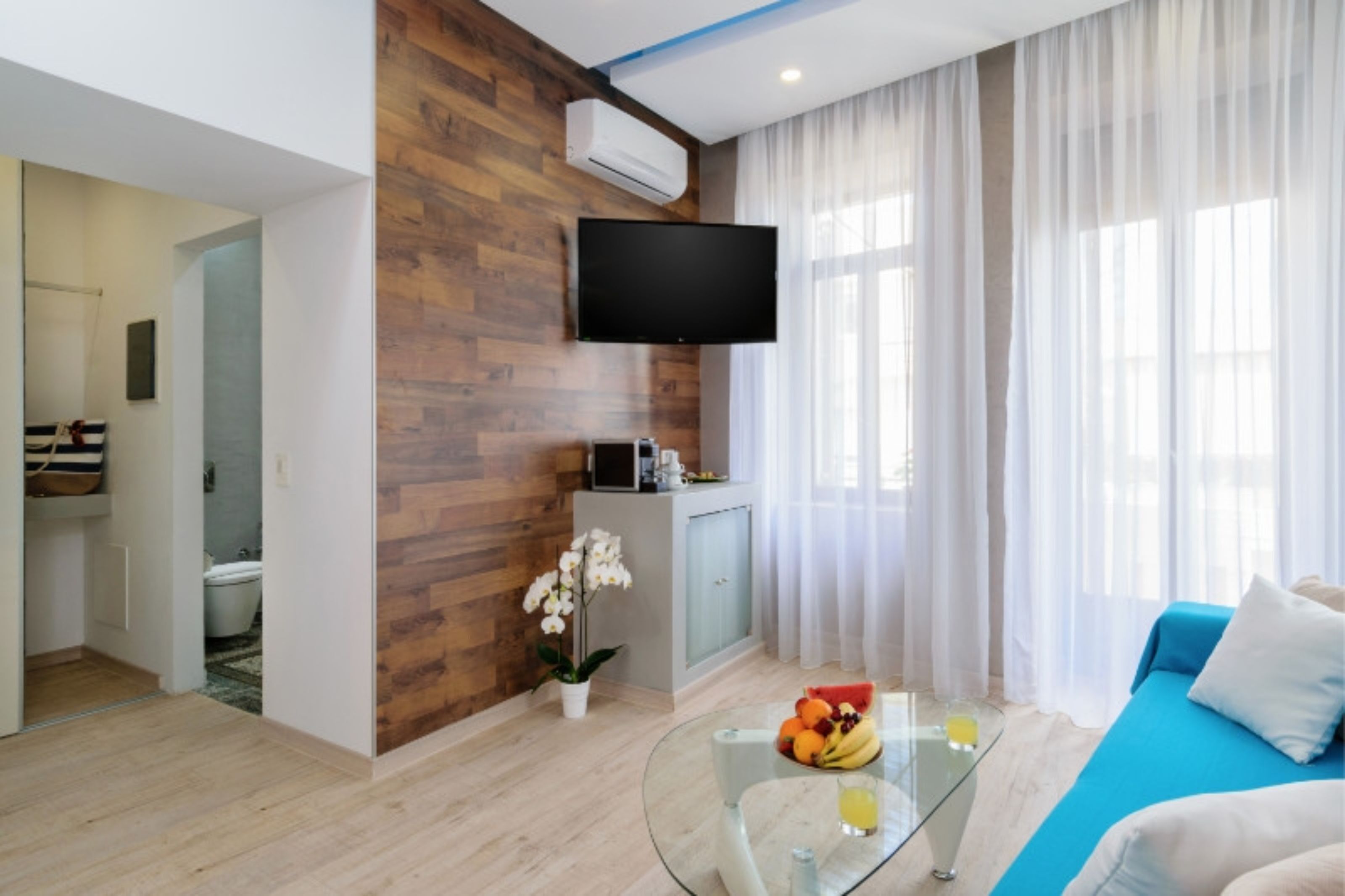 A modern living room with a wooden accent wall, a flat-screen TV mounted above a kitchen area, a glass coffee table with fruits and a drink, and a blue sofa with white cushions.