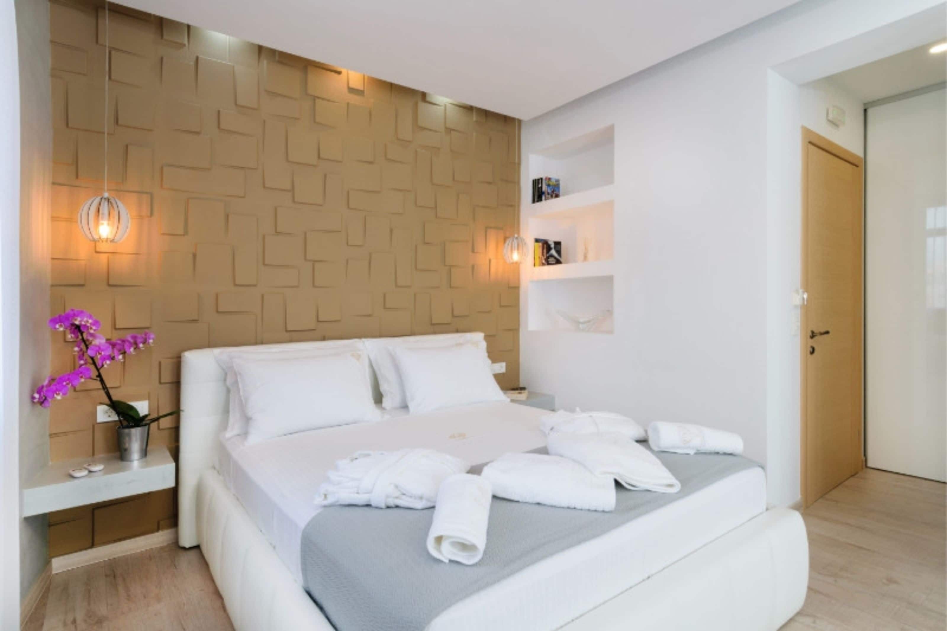 A modern hotel room with a neatly made bed, a bedside table with a lamp, a wall-mounted shelf with books, and a wooden accent wall.