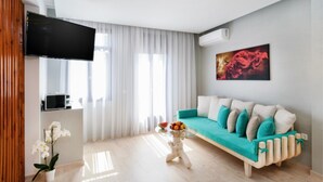 Een moderne woonkamer met een blauwgroene bank, witte kussens, een flatscreen televisie, een glazen salontafel met fruit, en een schilderij aan de muur.