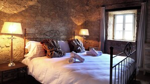 Une chambre rustique avec un mur en pierre, un lit recouvert de draps blancs, deux lampes de chevet et une fenêtre avec des rideaux.