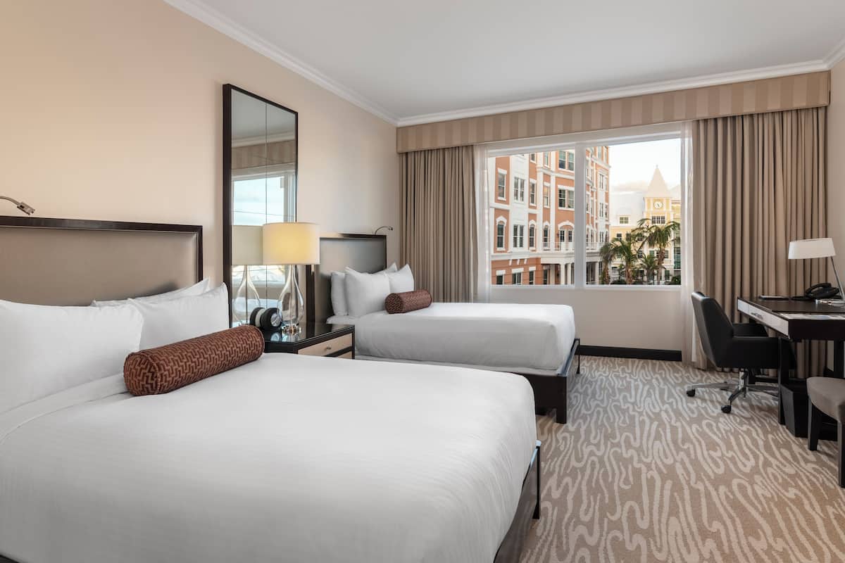 Ein Hotelzimmer mit zwei Betten, einem Schreibtisch, einem Stuhl, einer Lampe und einem großen Fenster mit Vorhängen.