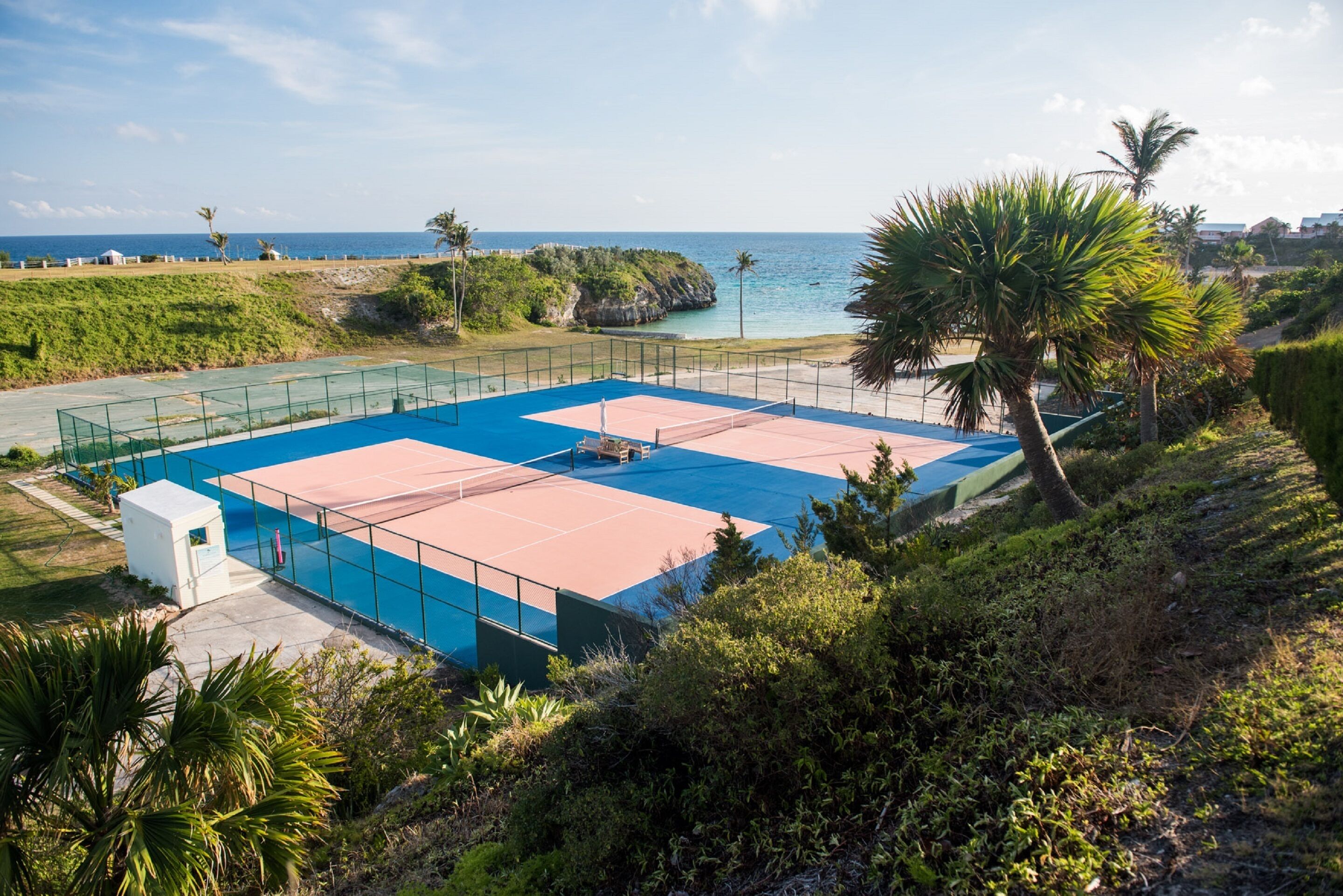 Una cancha de tenis con red, rodeada de vegetación y vista al océano.