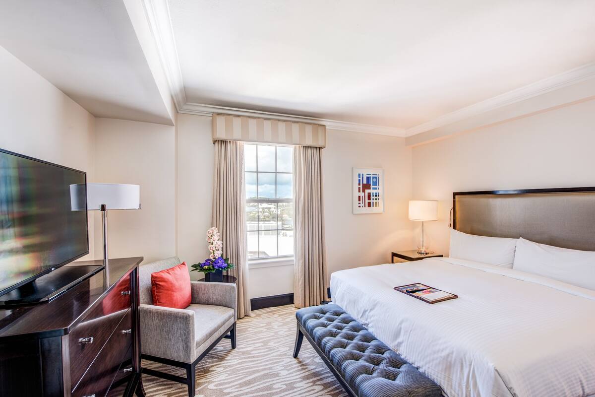 Ein Hotelzimmer mit einem großen Bett, einem Fernseher, einem Schreibtisch mit Schubladen, einem Stuhl, einer Bank, einem Fenster mit Vorhängen und einem Nachttisch mit einer Lampe.