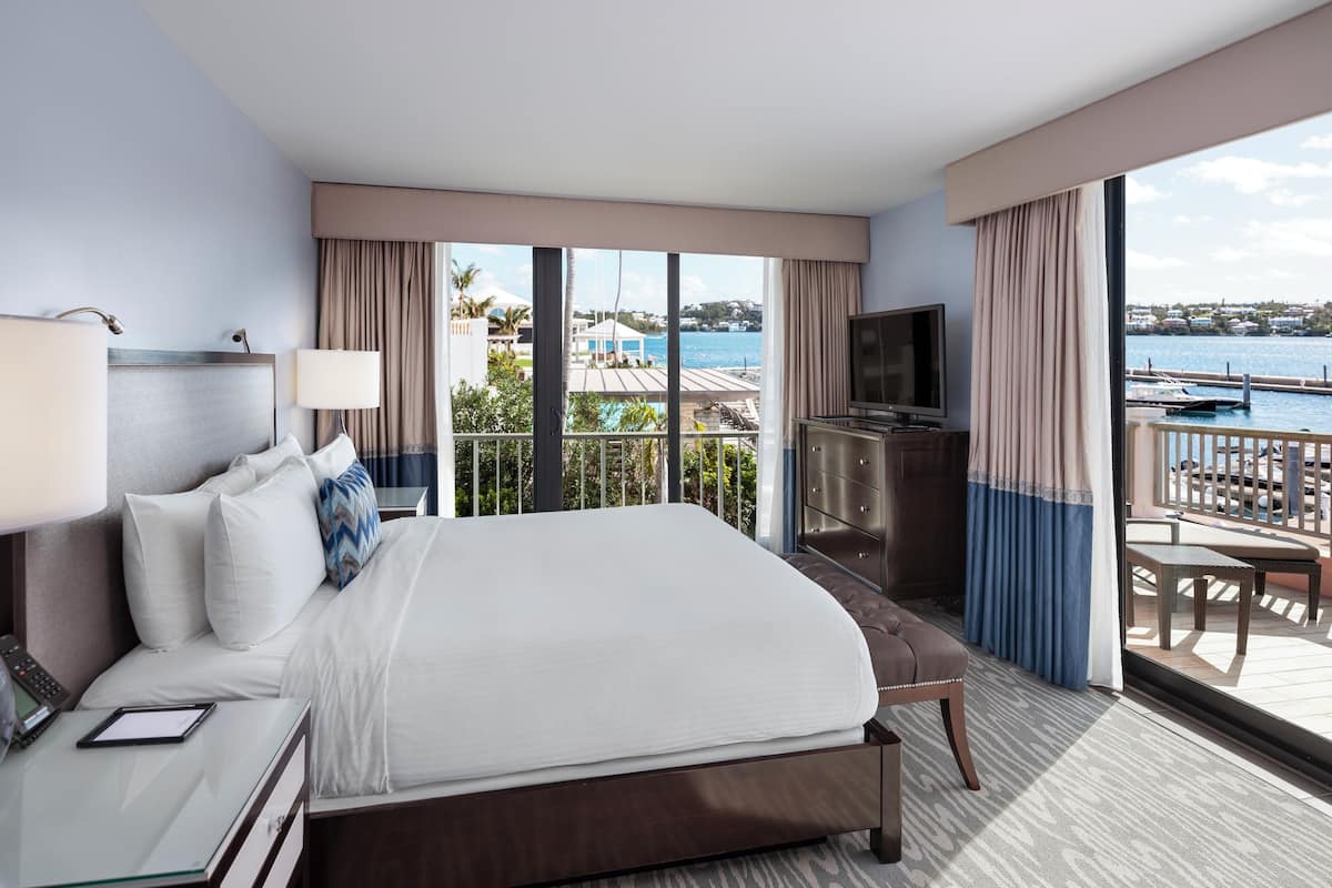 Ein Hotelzimmer mit einem großen Bett, einem Fernseher, einem Schreibtisch mit Telefon und Blick auf das Meer und die Gebäude.