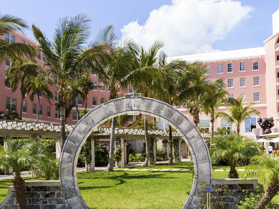Un arco de piedra circular en un jardín bien cuidado, con palmeras y un sendero pavimentado que lleva a un edificio de fachada rosa.