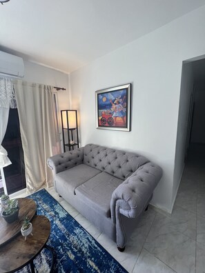 A living room with a grey sofa, a blue patterned rug, a framed painting on the wall, and a small table with potted plants.