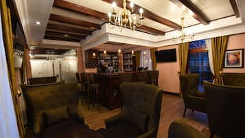 Zona bar con soffitto in legno, sgabelli da bar e scaffalatura per vini. In primo piano, due poltrone imbottite e un tavolo.