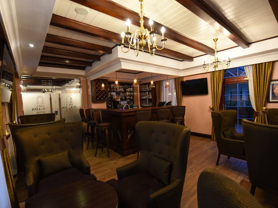 Zona bar con soffitto in legno, sgabelli da bar e scaffalatura per vini. In primo piano, due poltrone imbottite e un tavolo.