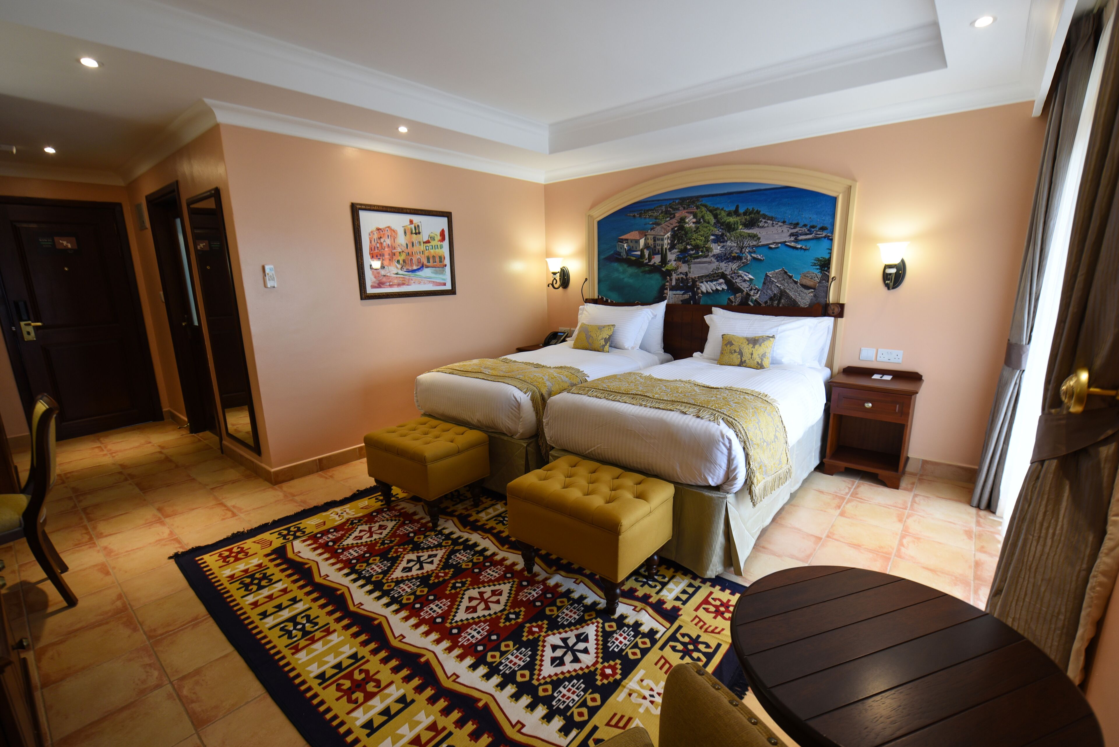 Habitación de hotel con una cama grande, dos mesitas de noche, una pequeña mesa, una silla y una alfombra estampada.