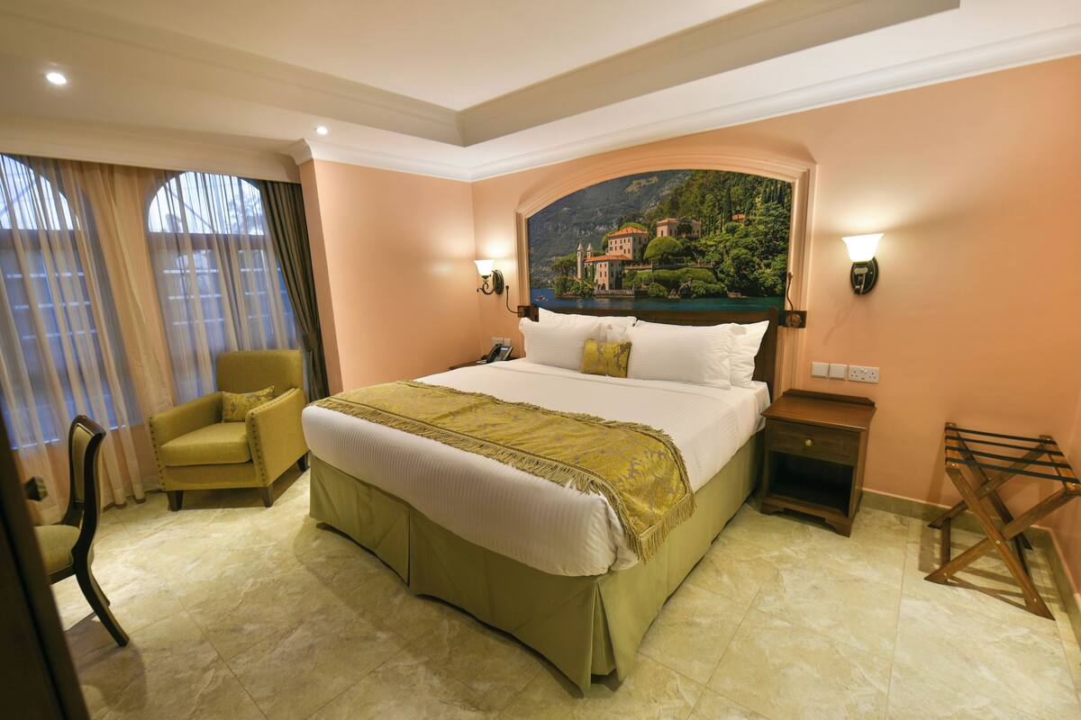 Habitación de hotel con una cama grande, una silla, una mesita de noche y un cuadro de un paisaje.