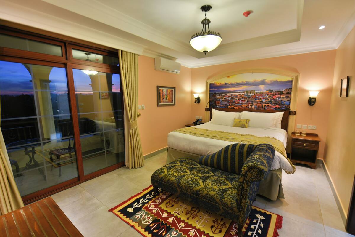 Habitación de hotel con una cama grande, una mesita de noche, una silla, un balcón con vistas y un cuadro en la pared.