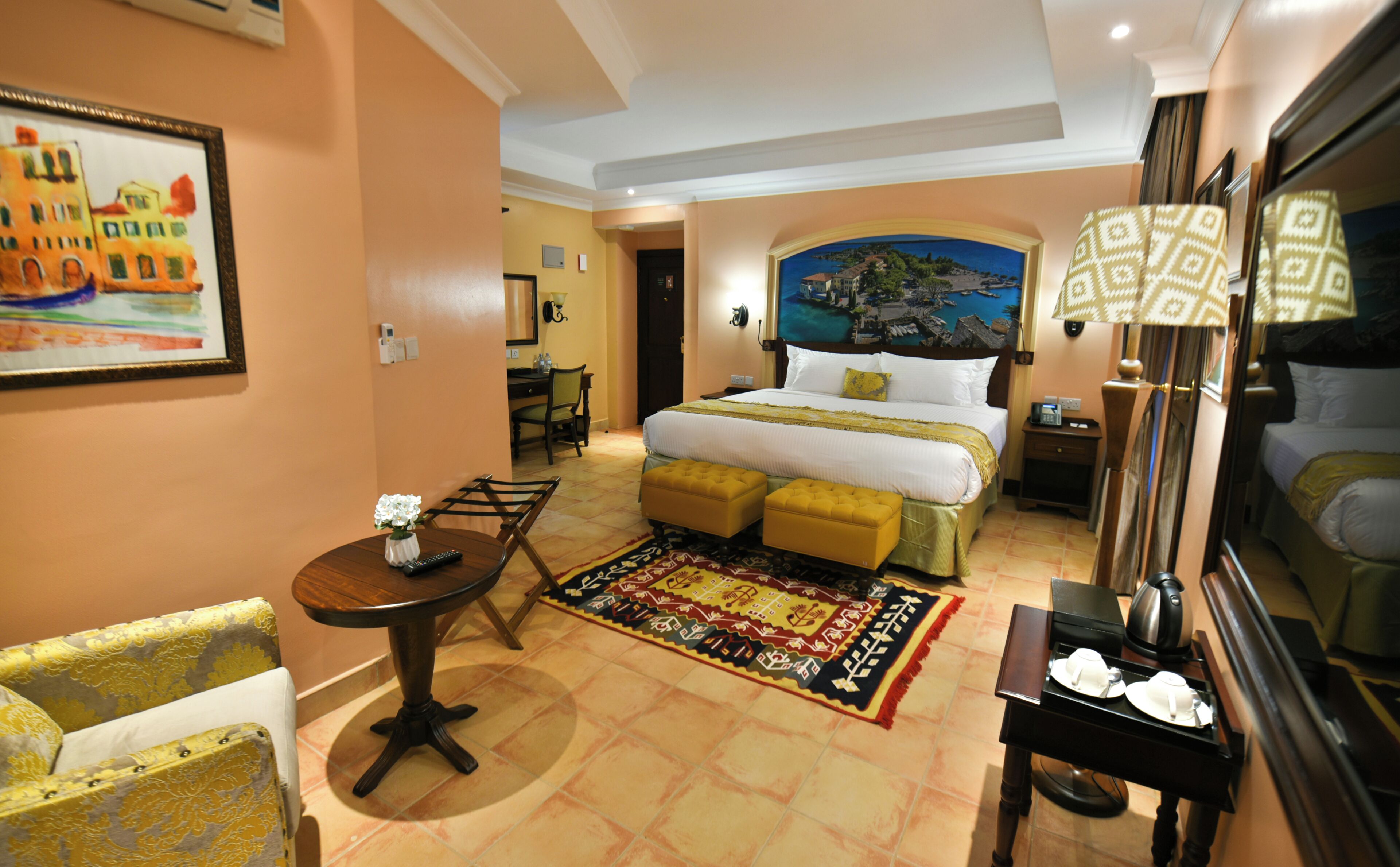 A hotel room with a bed, a desk, a chair, a table with a vase of flowers, and a painting on the wall.