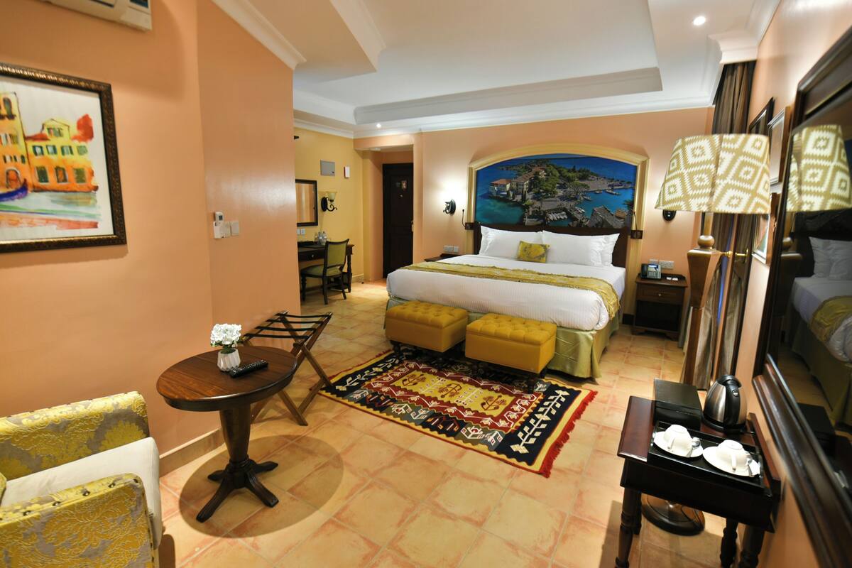 Habitación de hotel con una cama, un escritorio, una silla, una mesa con un jarrón de flores y un cuadro en la pared.