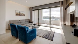 Una sala moderna con un sofá gris, dos sillones azules, una mesita de centro y un amplio ventanal corredero que da a un balcón con vistas a la ciudad.