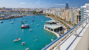 A marina with numerous boats, a modern building with a glass facade, and a cityscape in the background. - Superlative Penthouse, Valletta and Harbour Views (Sliema)