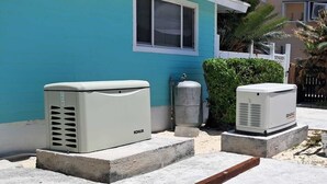 Deux unités de climatisation sont installées sur une terrasse en béton devant une maison à la façade bleue.