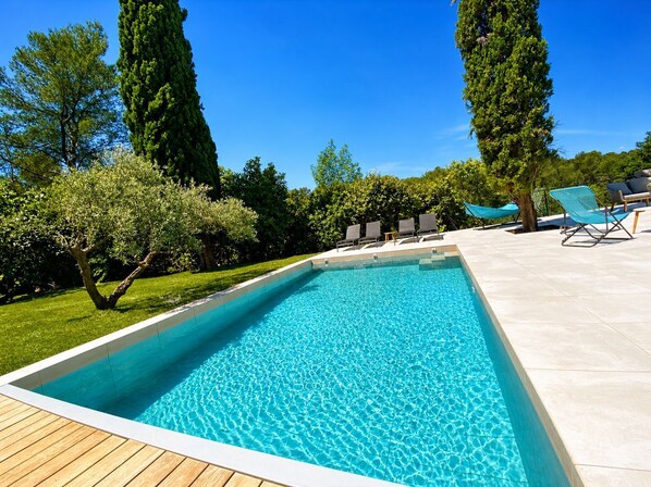 A rectangular swimming pool with clear blue water, surrounded by a wooden deck and a tiled patio area with lounge chairs.