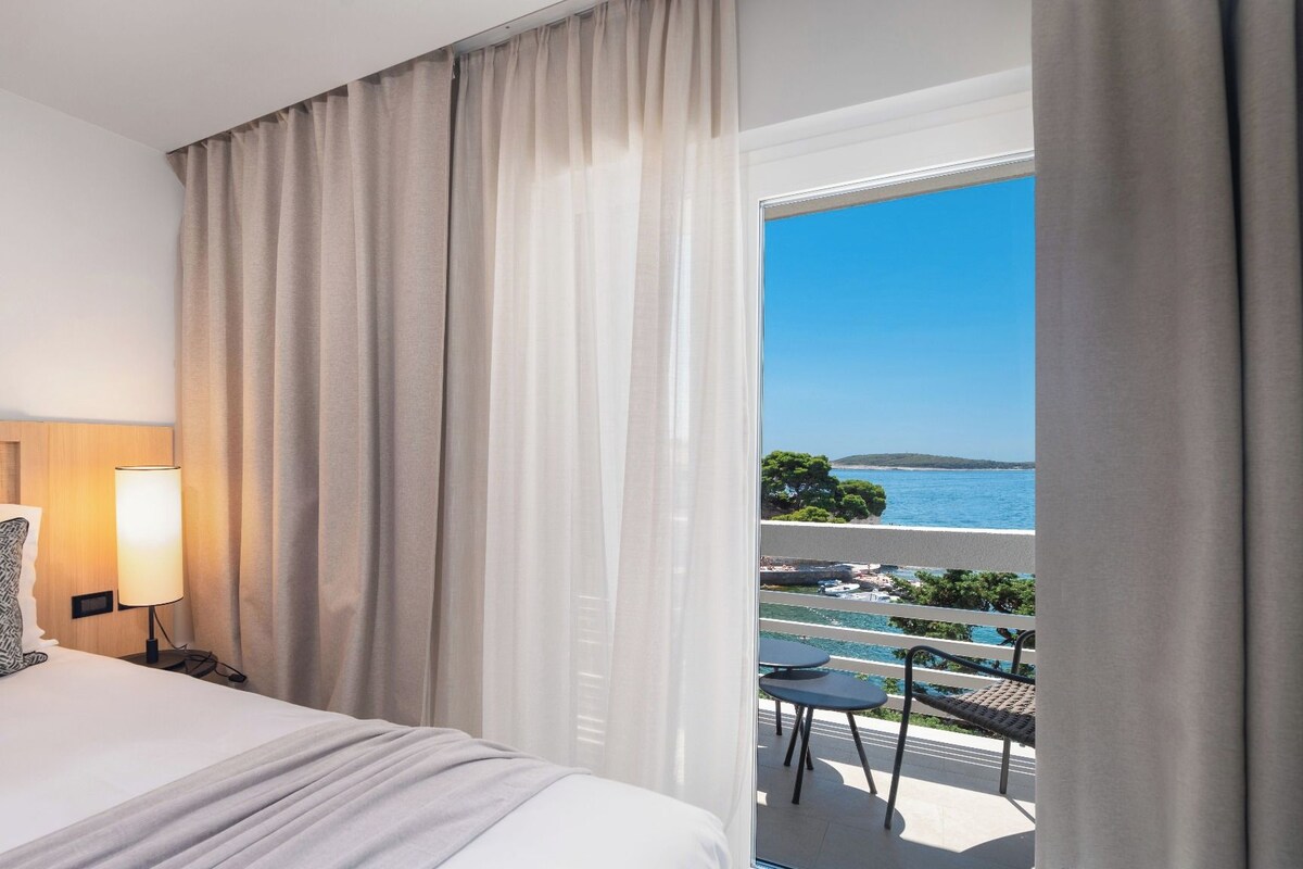 A hotel room with a bed, a bedside lamp, a view of the sea, and a balcony with outdoor furniture.