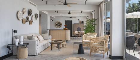Una sala moderna con sofá, mesa de centro y sillas de mimbre. Hay una zona de comedor con mesa y sillas, y un ventanal con vistas al exterior.