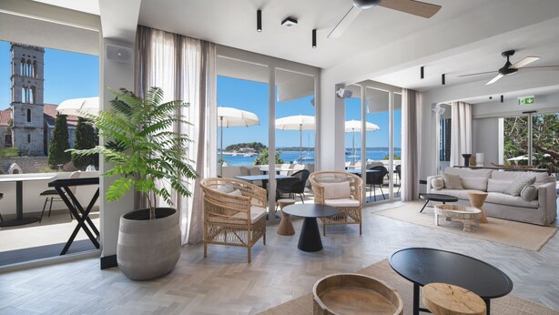 A spacious living area with a large window offering a view of a historic tower and the sea. There are wicker chairs, a sofa, and a coffee table. - Beach Bay Hvar Hotel (Hvar)