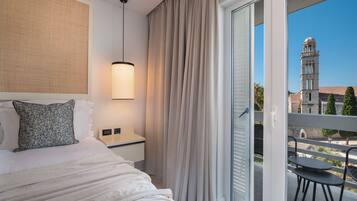 A hotel room with a bed, a bedside table, a lamp, a view of a balcony with a chair, and a building with a tower in the background.