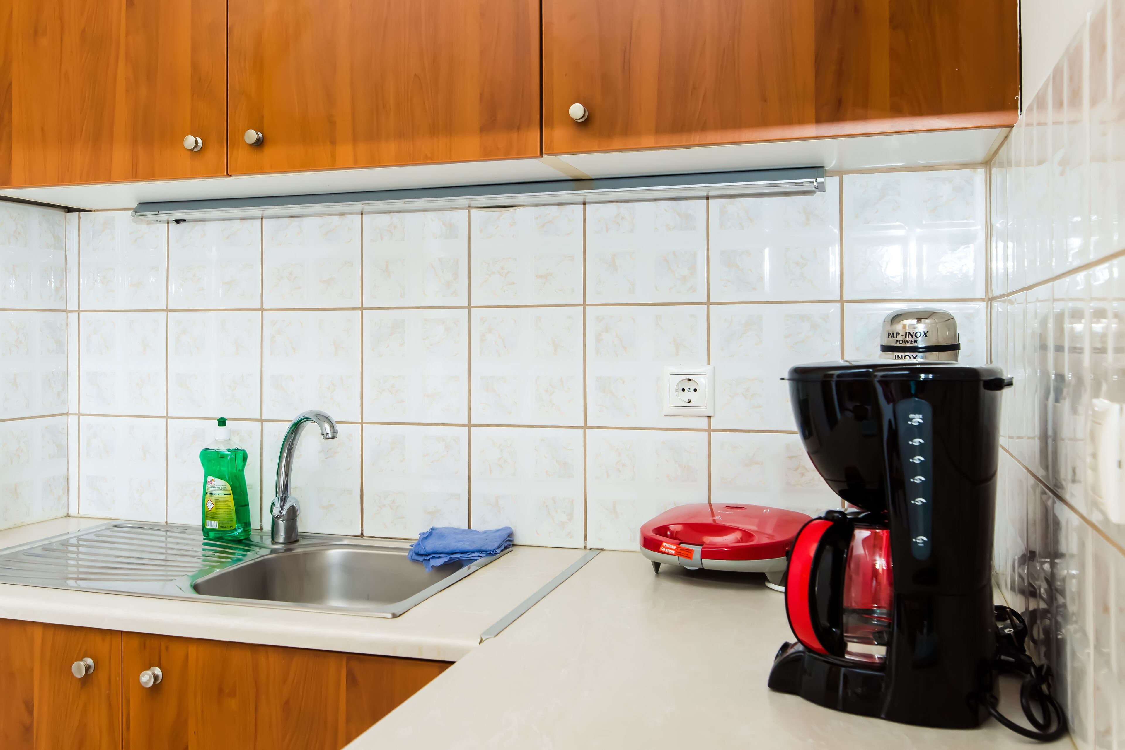 Een keuken met houten kastjes, een roestvrijstalen spoelbak, een fles afwasmiddel, een blauwe doek en een rode koffiezetapparaat.