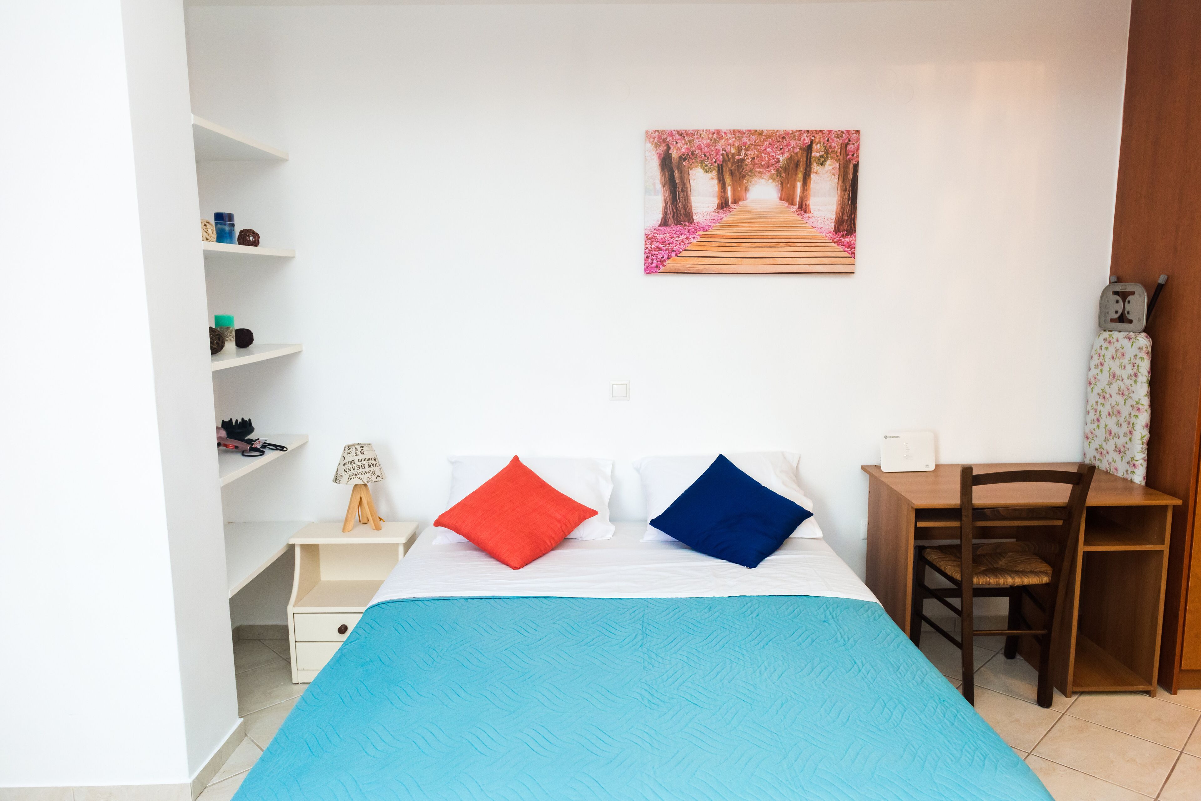 Een slaapkamer met een bed, een bureau, een stoel, een plank met spullen en een schilderij aan de muur.