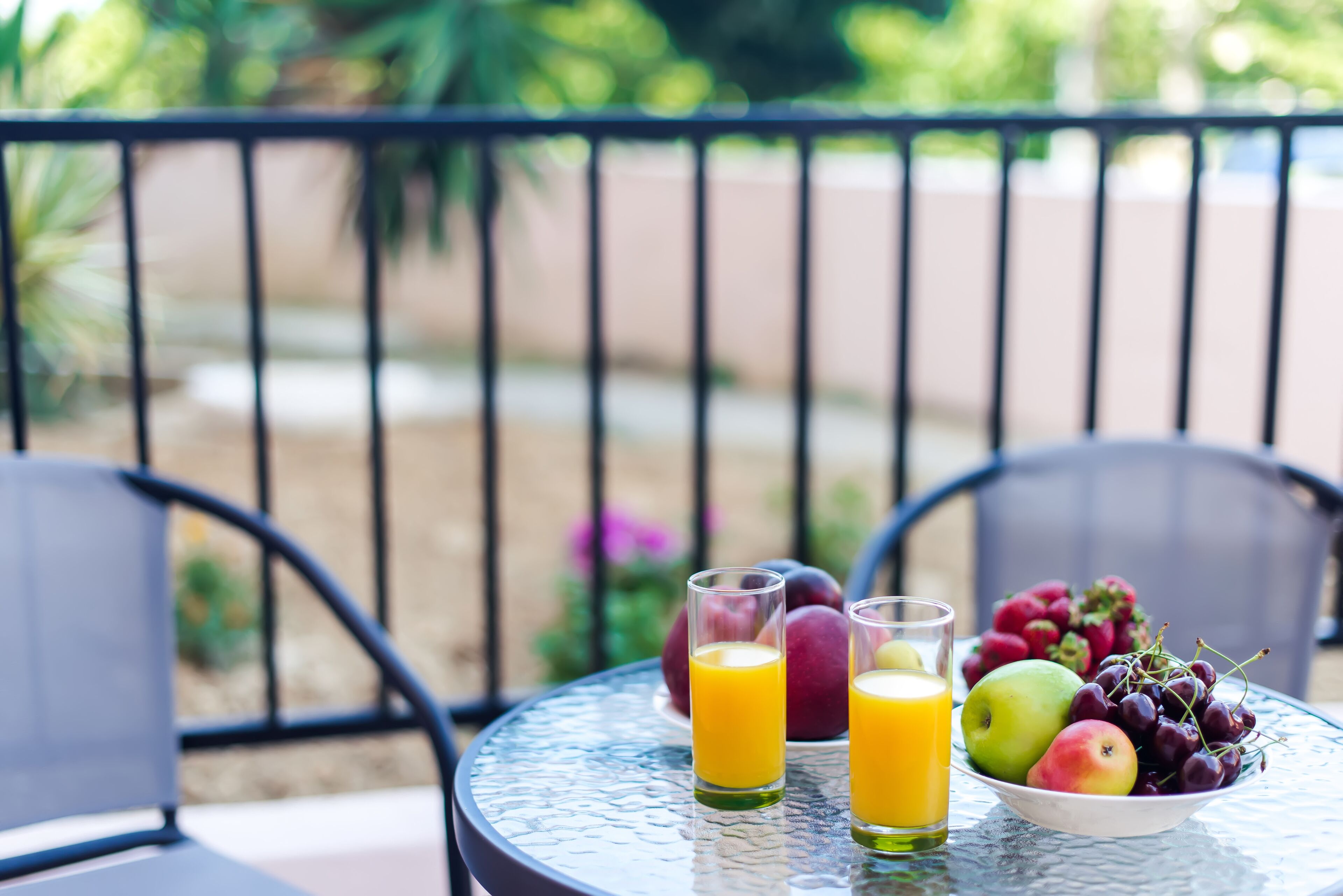 A table with a glass of orange juice, a glass of juice, and a bowl of fruit on a balcony.