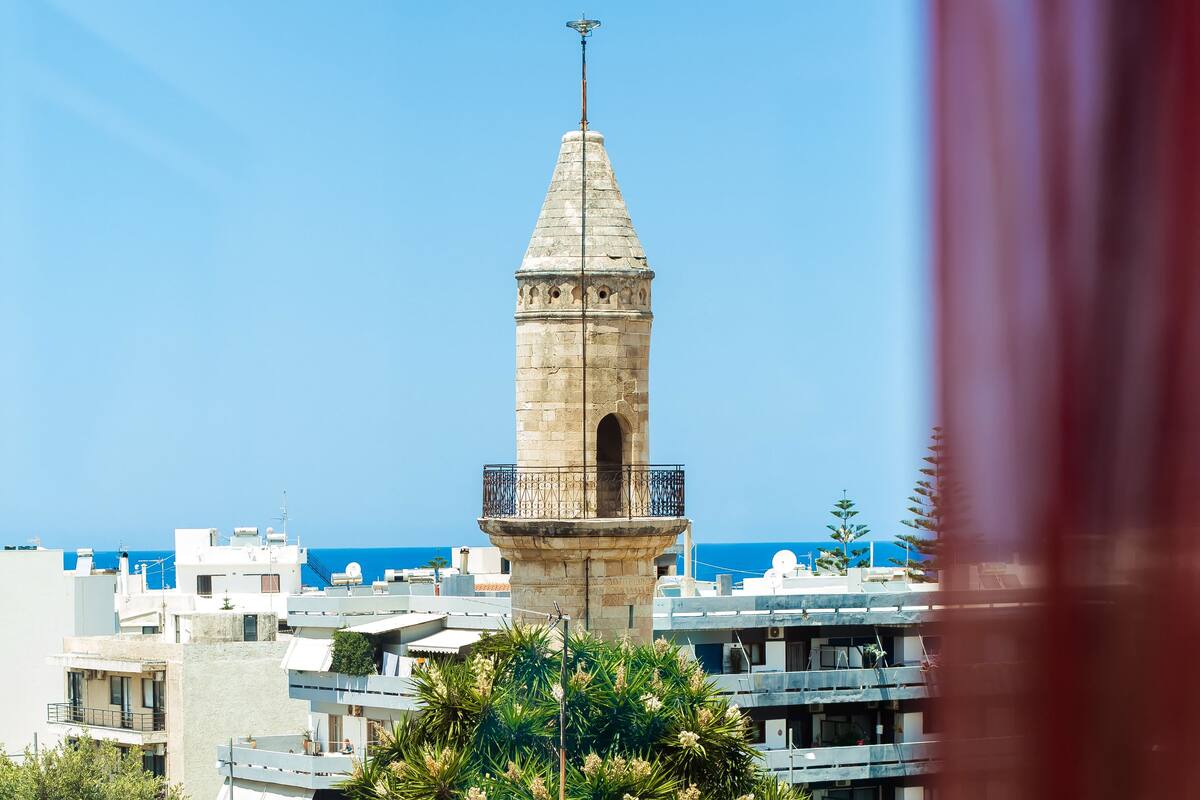 Una torre con techo puntiagudo y un balcón, rodeada de edificios y un cielo despejado.
