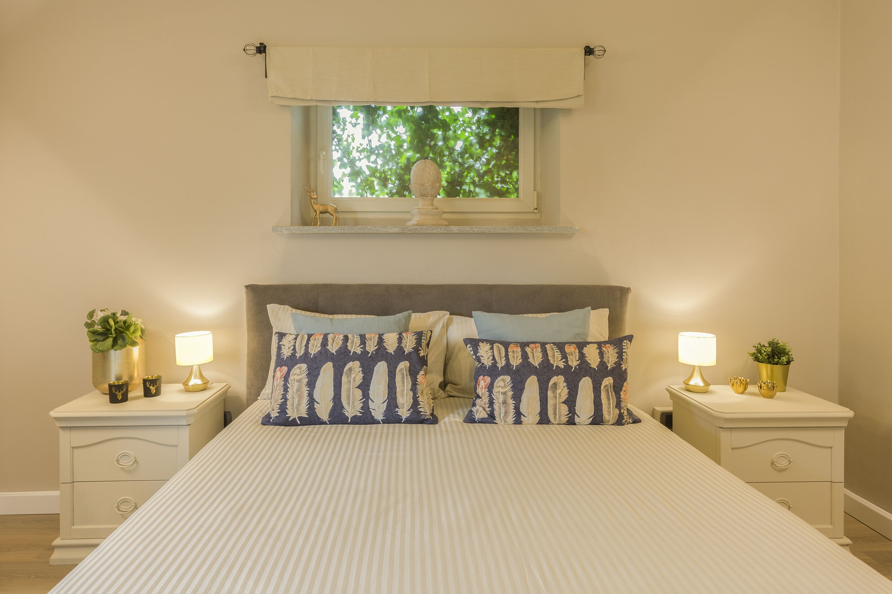 Une chambre avec un grand lit, deux tables de chevet avec des lampes, une fenêtre donnant sur des arbres et un objet décoratif posé sur le rebord de la fenêtre.