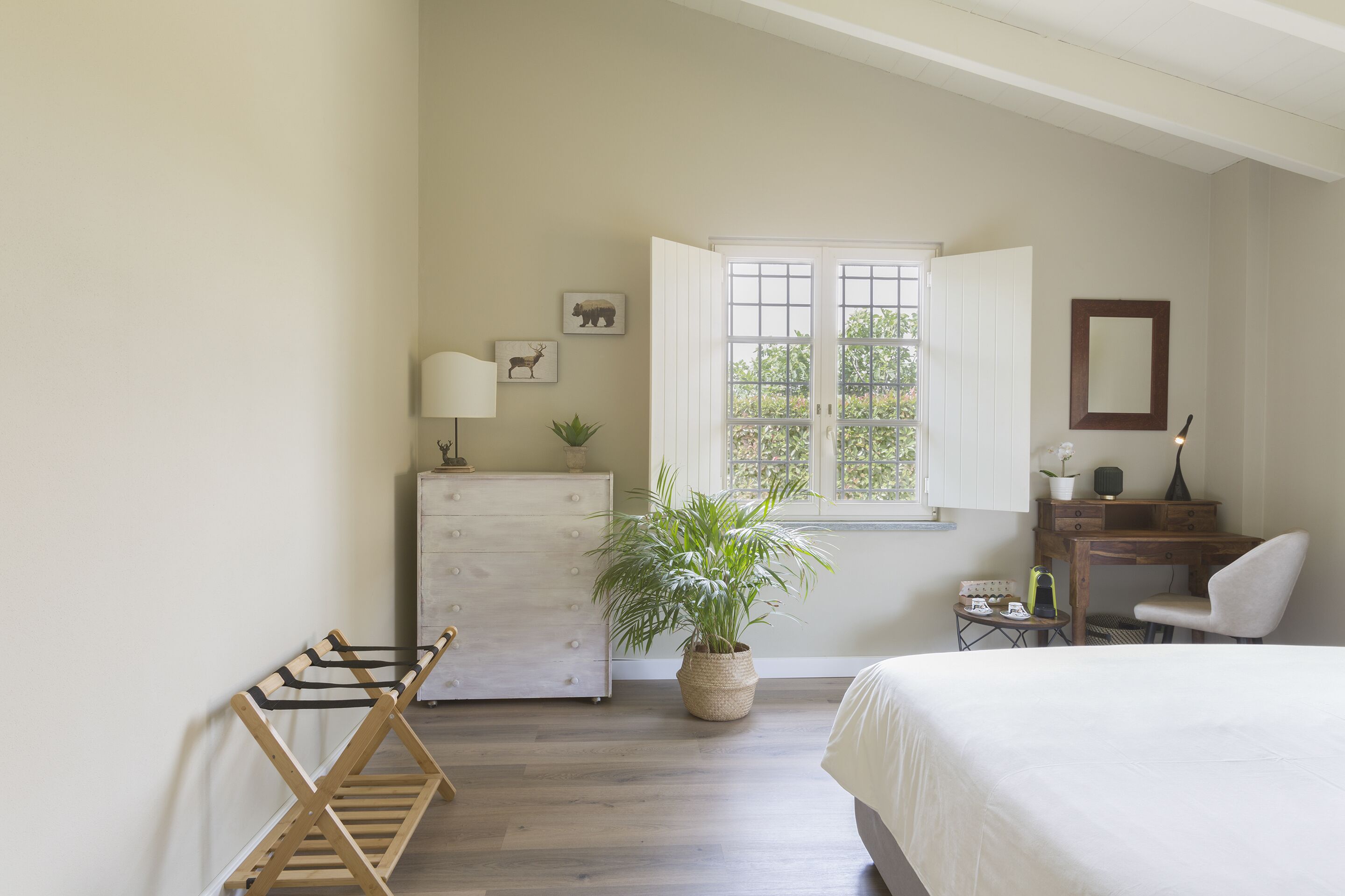 Ein Schlafzimmer mit einem Bett, einem Schreibtisch, einem Stuhl, einer Kommode, einer Lampe, einer Topfpflanze und einem Fenster mit geöffneten Klappläden.
