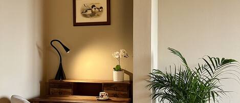 A corner room with a wooden desk, a chair, a framed picture on the wall, a lamp, a small potted plant, and a large potted plant.