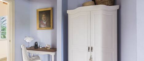 Una habitación con una silla blanca, una mesita con una lámpara, un retrato enmarcado y un armario blanco.