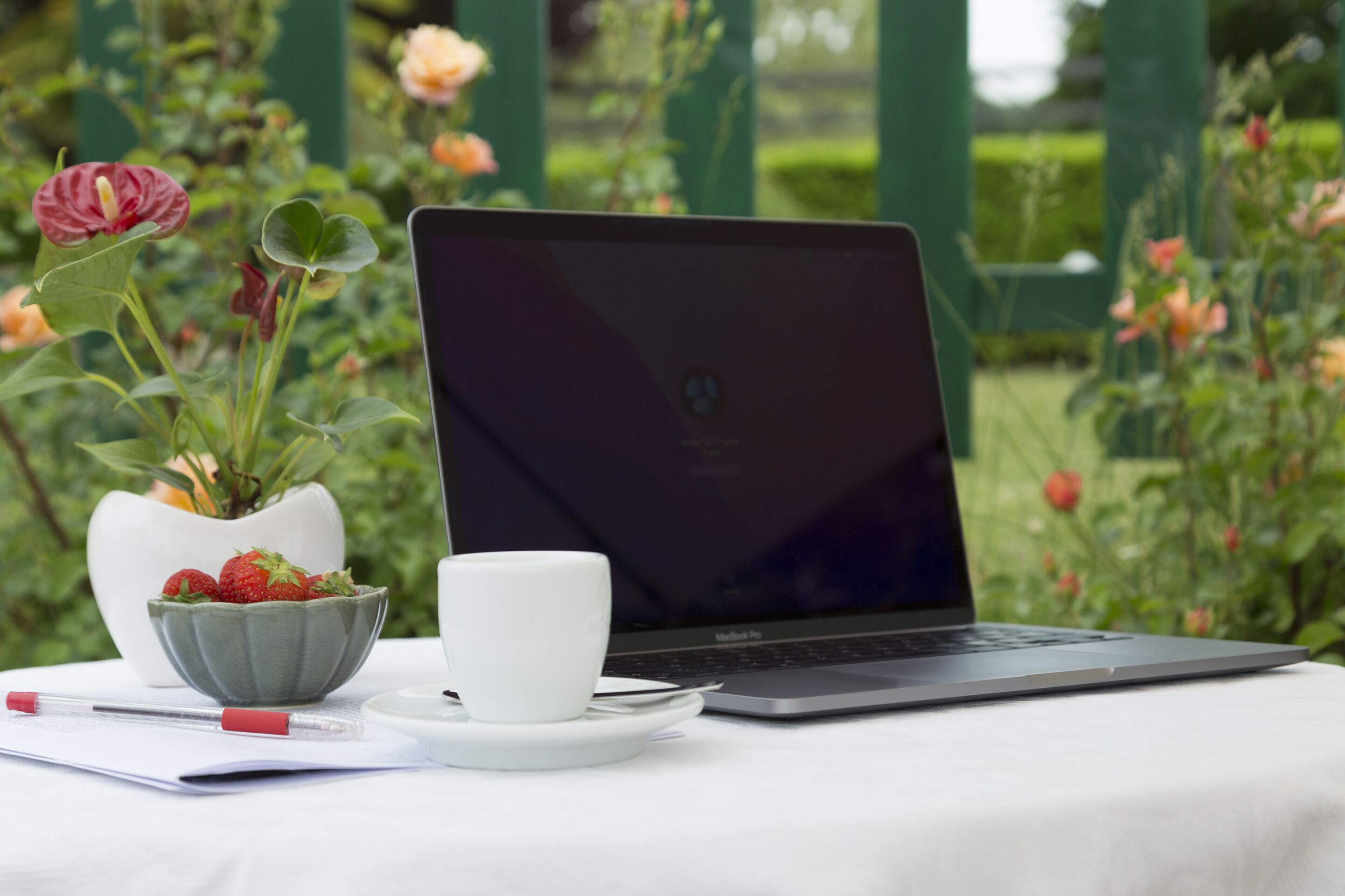 A laptop on a table with a white cup and a bowl of strawberries.