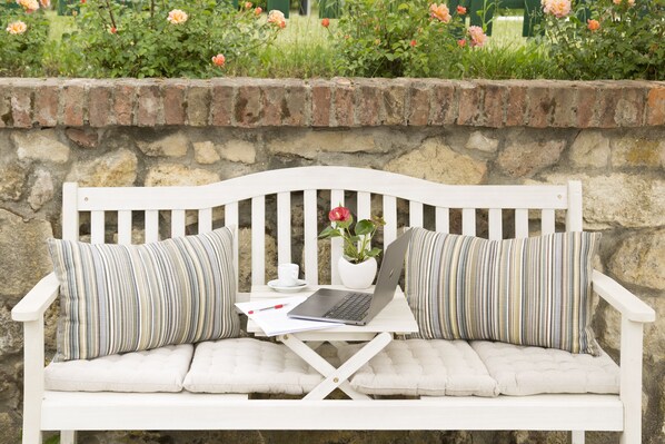 Un banc blanc, sur lequel sont posés un ordinateur portable et une tasse, entouré d’un mur de pierre et de fleurs en pleine floraison.